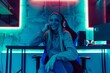 © Svetlana - The young blonde woman radiates confidence as she smiles while sitting comfortably at a table in her lively gaming room, surrounded by colorful LED lights and gaming gear.