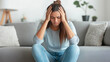 © MargoArt - Young woman sitting on sofa with her head in her hands, looking stressed and overwhelmed, conveying emotions of frustration and anxiety in home environment