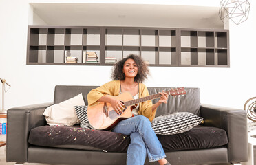 A happy, talented Black mixed-race woman is playing acoustic guitar and singing a song on a sofa in a domestic house. A beautiful female artist is practicing her singing skills before a performance.