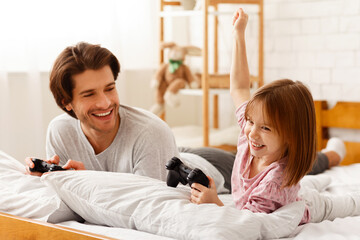 Wall Mural - Cheerful family father and little daughter playing videogame, laying on bed in bedroom at home