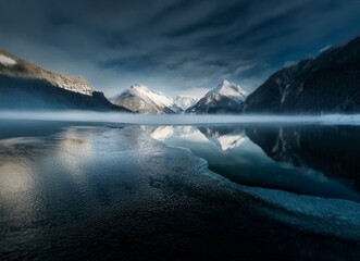 Poster - A calm, still lake reflects snow capped mountains under a dark sky.