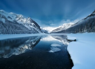 Poster - A mountain lake in the winter with a blue sky, snow covered mountains and trees reflected in the still water.
