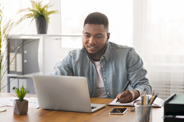 Poster - Young black businessman using laptop and taking notes at worklace in modern office,free space