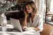 © sergign - A woman in white blouse working on a laptop in a cozy cafe with elegant decor and natural light coming through windows.