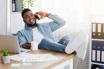Poster - Joyful black manager talking on phone and drinking coffee while having break at work, leaning back on chair and putting legs on table, copy space