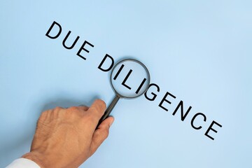 A hand holding a magnifying glass over the words “DUE DILIGENCE” on blue background. Business, finance, law, research concept.