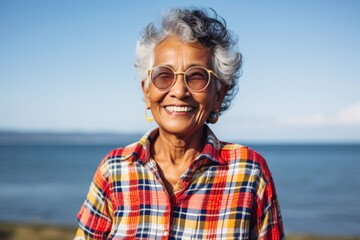 Sticker - Portrait of a grinning indian woman in her 80s dressed in a relaxed flannel shirt while standing against serene seaside background