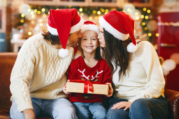 Cheerful little girl with xmas gift kissed by parents in santa hats over decorated kitchen background