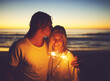 © StarDweller/peopleimages.com - Couple, love and sparkler on beach at night for new year celebration, party event and kissing on holiday. Tourist, people and fireworks at ocean sunset for vacation, travel and romance with affection