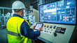 © Manatphon - A man in a hard hat and safety vest operates a digital control panel in an industrial setting, monitoring data on a large screen.