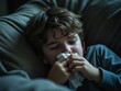 © Bianca - Sick child resting on a couch, holding a tissue to his nose, suffering from flu or cold symptoms in a dimly lit indoor setting, representing illness and recovery in children