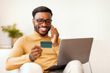 Poster - Online shopping concept. Happy afro man talking on cellphone using credit card and laptop sitting on sofa indoor. Selective focus