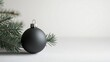 © Studio 54 - A single Christmas tree ornament with matte black finish and pine branches. Modern and minimalistic design with soft white backdrop and copy space.