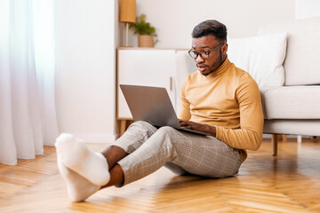 Aufkleber - Shocked african american man using computer reading news on laptop sitting on floor at home. Selective focus