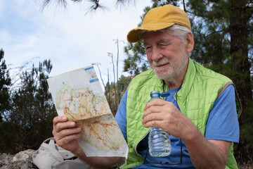 Senior carefree bearded man in outdoor excursion in the forest sitting holding a water bottle while looks at the map of footpaths. Elderly male enjoys healthy lifestyle and activity