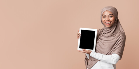 Poster - Cheerful afro islamic girl in headscarf presenting digital tablet with empty black screen, standing with device over yellow studio background, mockup