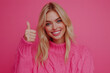 © ALL YOU NEED studio - beautiful blue eyed blonde young woman wearing knited vibrant pink sweather is smiling and showing thumb up on camera, isolated on pastel solid smooth pink background, web banner with copy space