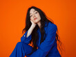 © Amy - Confident woman in bold blue pantsuit sitting against vibrant orange background, modern and trendy fashion portrait