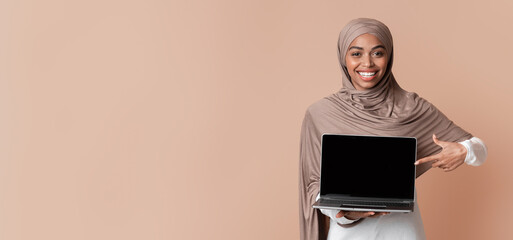 Poster - New Website. Smiling Black Muslim Girl In Hijab Holding Laptop Computer With Blank Black Screen For Mockup And Pointing At It, Yellow Background