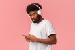 © Prostock-studio - Nice music app for smartphone. Young black man with wireless headset using phone over pink background