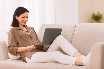 Poster - Happy Lady. Excited entrepreneur woman wearing brown blouse working on laptop sitting on a couch at home, copyspace