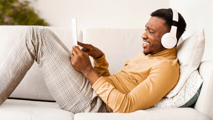 Aufkleber - Weekend Concept. Happy African American Guy In Headphones Using Tablet Computer Listening To Music Relaxing Lying On Couch Indoor