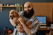 © Seventyfour - Portrait of man with beard holding cute fluffy dog in relaxed modern office setting with wooden accent wall and computer in background, smiling warmly and engaging camera