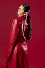 Wall Mural - Fashion asian brunette in red leather coat standing with hand on hip against red background, hair in ponytail