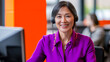 © Ron - Senior Asian woman in a bright purple blouse wearing a headset, smiling warmly while assisting a client at her desk in a modern office with bold orange accents.