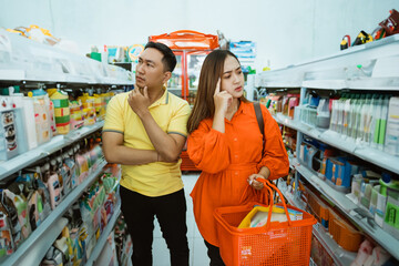 asian man and woman with thinking expression will take goods on shelf while shopping carrying cart in minimarket