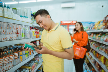 young man holding goods and mobile phone while standing shopping at minimarket