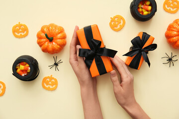 Wall Mural - Female hands with Halloween gifts, pumpkin, spiders and pots with candies on beige background. Top view