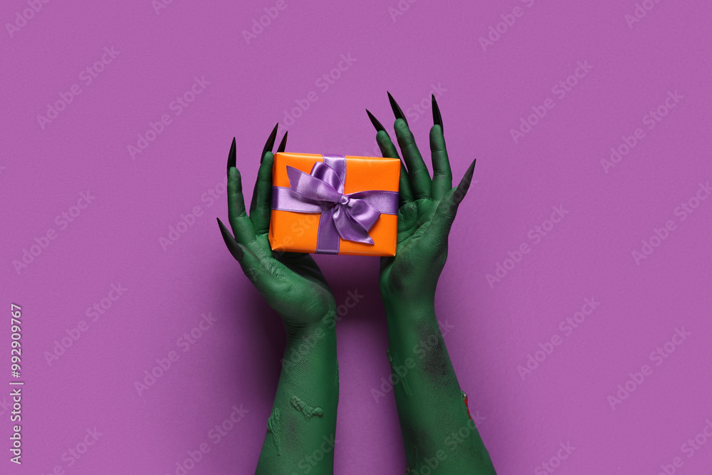 Green hands of witch with claws and gift box on purple background. Halloween celebration