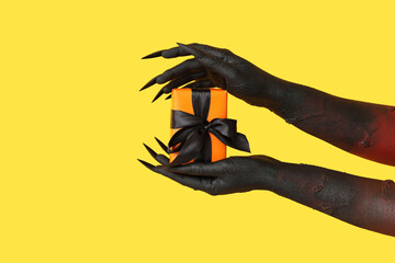 Black hands of witch with gift box on yellow background. Halloween celebration