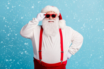 Wall Mural - Photo of pensioner old man grey beard hold specs hand hip self-assured prepare check sledge wear santa costume red suspenders spectacles white gloves headwear isolated blue color background