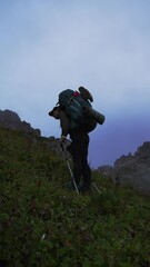 a lone hiker struggling to ascend a steep, grassy mountain slope at dusk, carrying a large backpack 