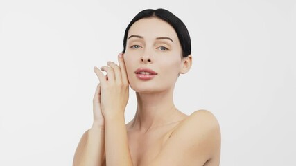 Wall Mural - A young woman in a studio against a white background gently touches her flawless skin. Her expression conveys joy and harmony, highlighting her allure and well-groomed appearance.