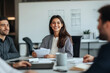 © gfx_yasir - A woman smiles at the camera, seated in a meeting with two other people