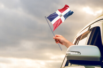 Waving the Dominican Republic flag against the sunrise or sunset from a car driving along a country road. Holding the Dominican Republic flag, traveling by car, on a weekend trip.