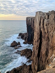 Wall Mural - A high cliff overlooking a vast ocean with crashing waves against the rocks in the foreground.