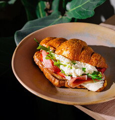 Delicious croissant sandwich with prosciutto, soft cheese and arugula on a plate