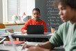 © Mediaphotos - Portrait of Black young woman using laptop in school classroom with defocused students in foreground, copy space