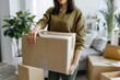 © LidiaLens - Young woman carrying a sealed cardboard box in a plant-filled living room, preparing for a move or reorganizing her home, surrounded by more boxes and cozy decor