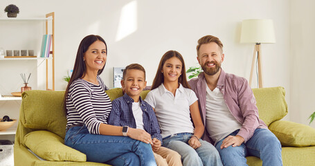 Smiling young happy family of four with two kids boy and girl sitting on the sofa at home and looking cheerful at camera. Portrait of parents with children. Family time and leisure concept.