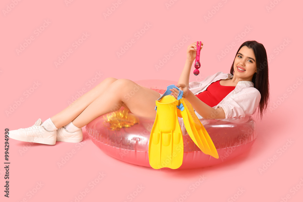 Young woman with inflatable ring, flippers and goggles lying on pink background