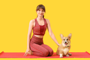 Wall Mural - Sporty young woman sitting on yoga mat with cute Corgi dog against yellow background