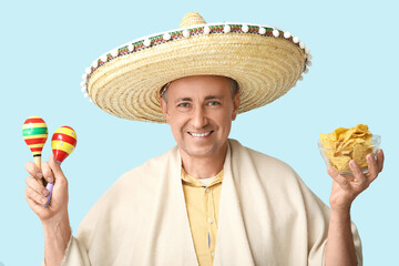 Wall Mural - Handsome mature Mexican man in sombrero hat with maracas and bowl of tasty nachos on blue background