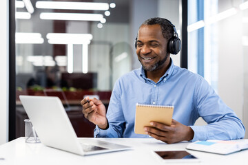 confident businessman with headphones participates in virtual meeting using laptop and taking notes.