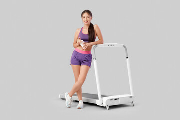 Wall Mural - Sporty Asian woman with water bottle and treadmill on light background