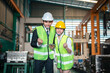 © Garun Studios - manufacturing, machinery, industrial, machine, blueprint, discussion, engineer, safety, foreman, hard hat. Two people in safety gear are standing in a factory. One of them is holding a tablet.